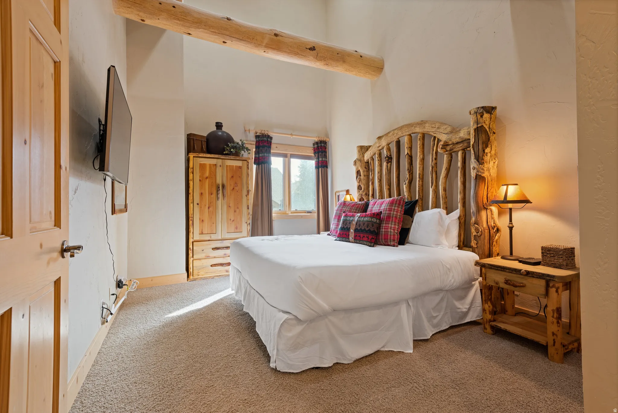 Spacious room featuring vaulted ceilings with exposed wood beams, a rustic wood-finish bed frame, light wood-finish door, and carpeted flooring