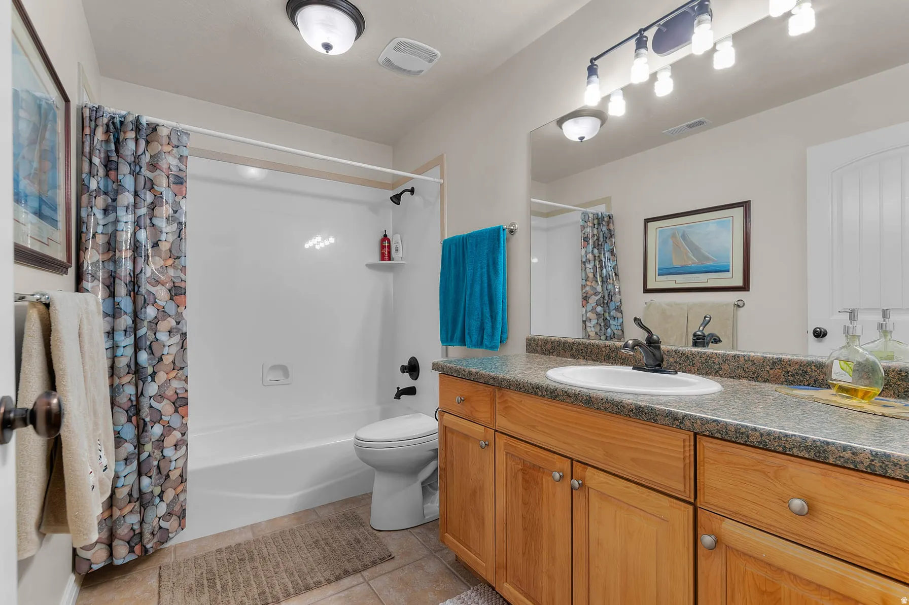 Bathroom featuring an integrated bathtub and shower, a single vanity with wood-finish cabinetry, a white basin, a stone-finish countertop, and tile flooring