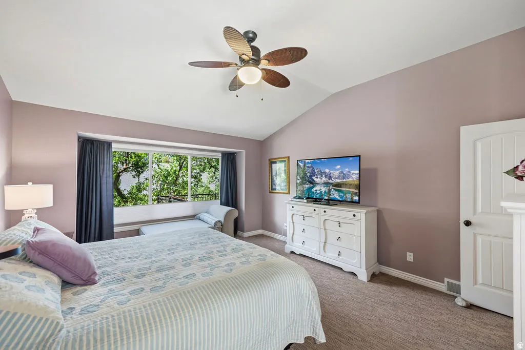 Vaulted ceiling bedroom featuring a wide window with exterior foliage, a ceiling fan with integrated lighting, and neutral-toned wall-to-wall carpeting