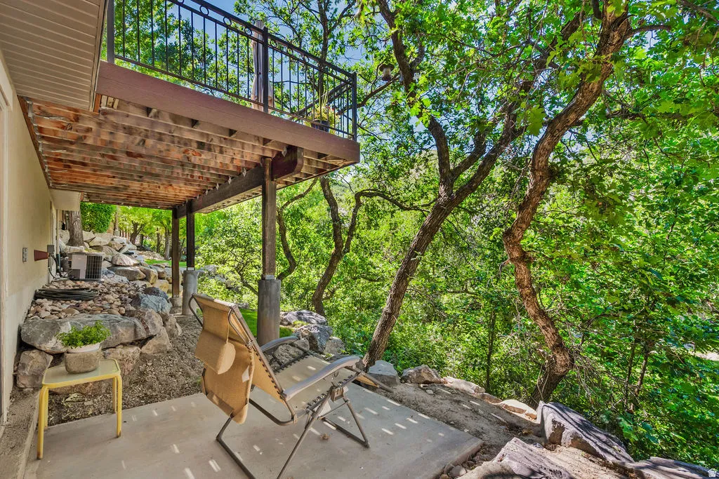 Tiered outdoor living spaces featuring a raised deck with black metal railings, a ground-level concrete patio, natural rock landscaping, and mature trees providing ample shade