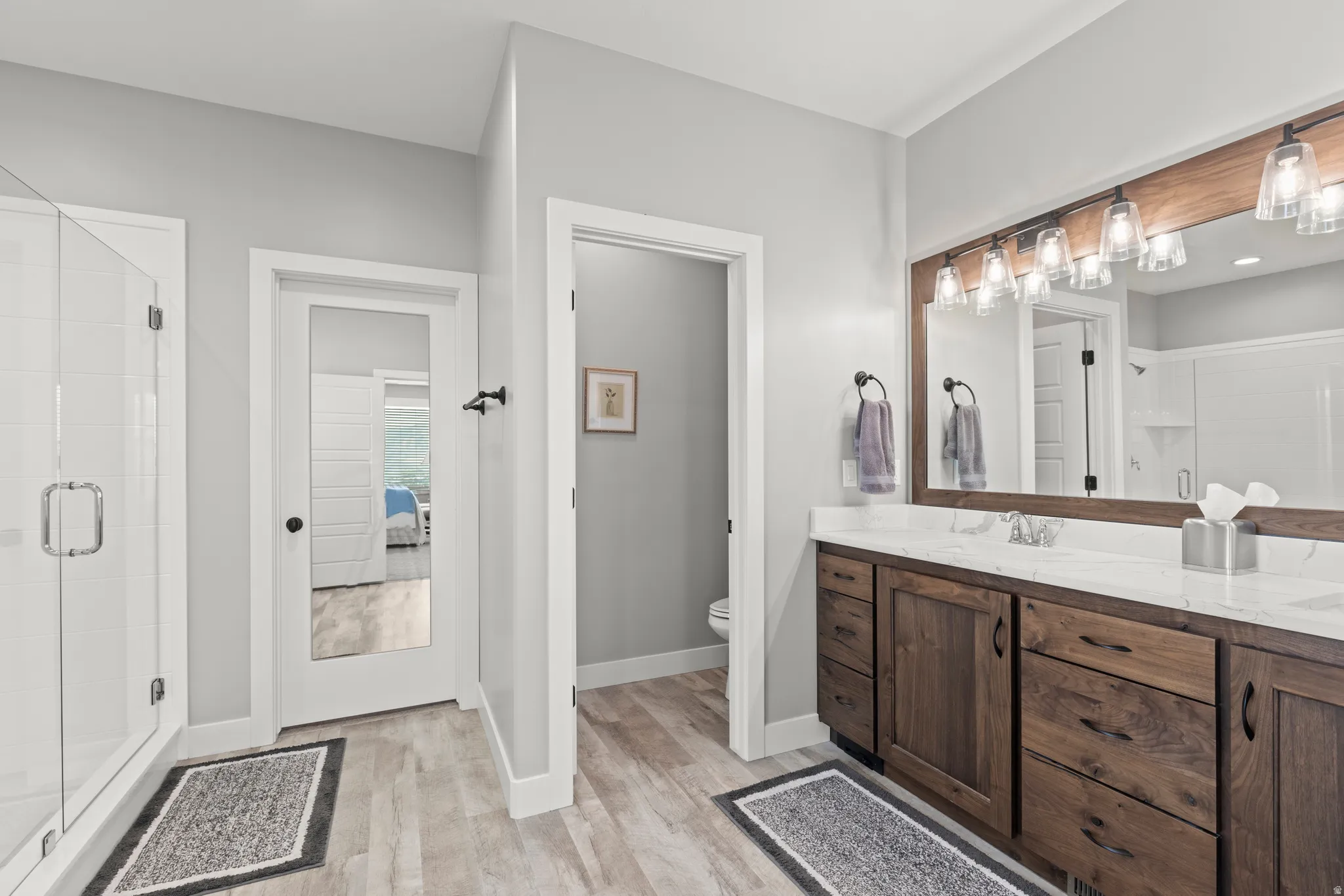 Bathroom featuring a double vanity with wood-finish cabinetry and light-toned countertops, a glass-enclosed shower, and wood-finish flooring