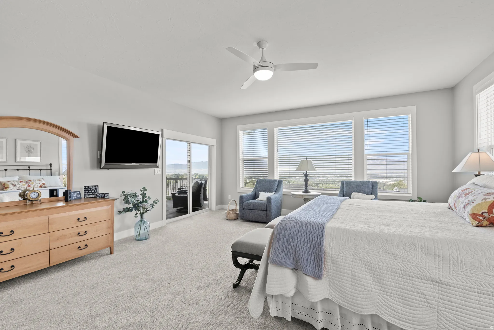 Spacious carpeted room featuring a ceiling fan with integrated lighting, a large window array with horizontal blinds, and a glass sliding door providing outdoor access