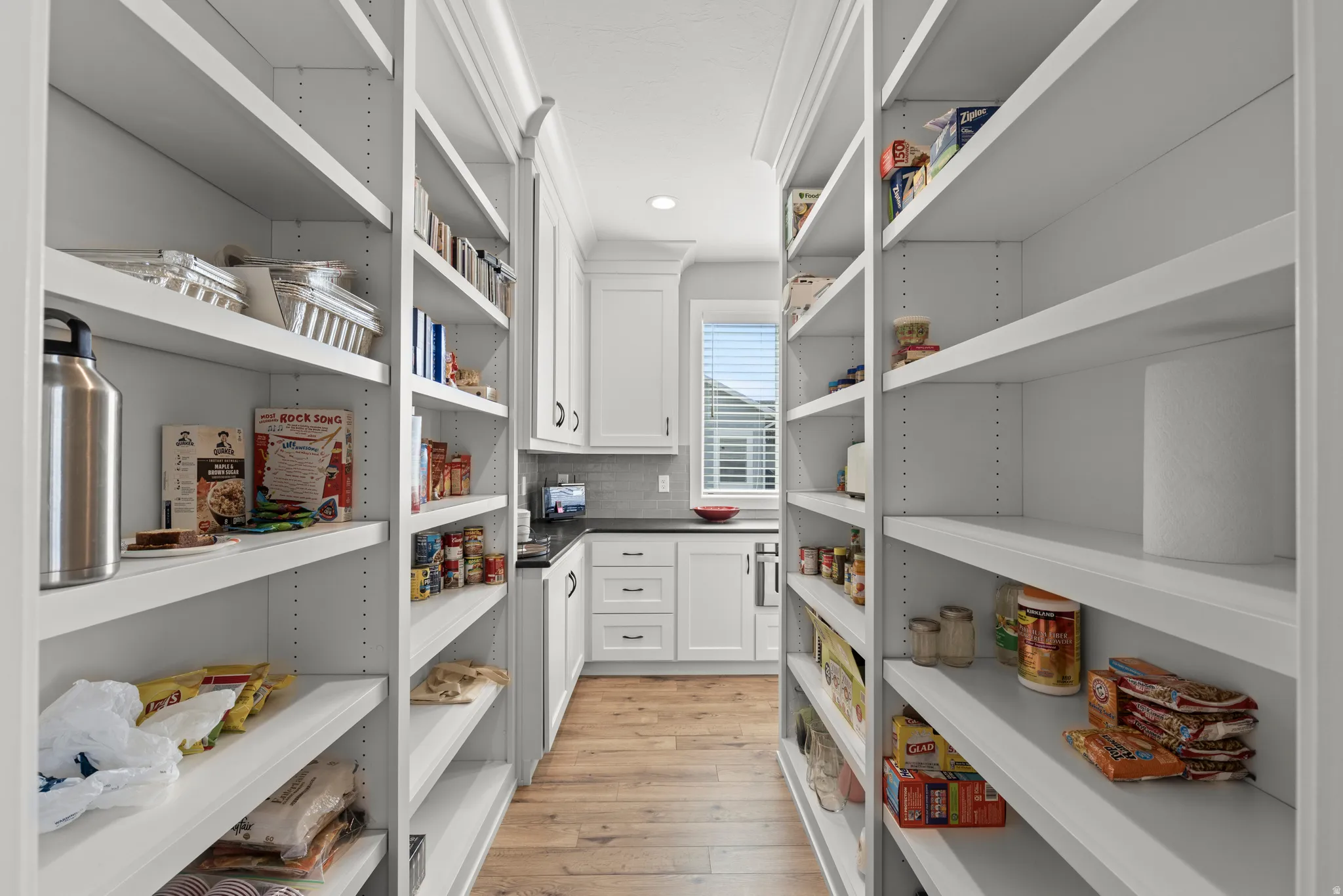 Walk-through pantry featuring extensive adjustable shelving, wood-finish flooring, white cabinetry, dark countertops, and a subway tile backsplash