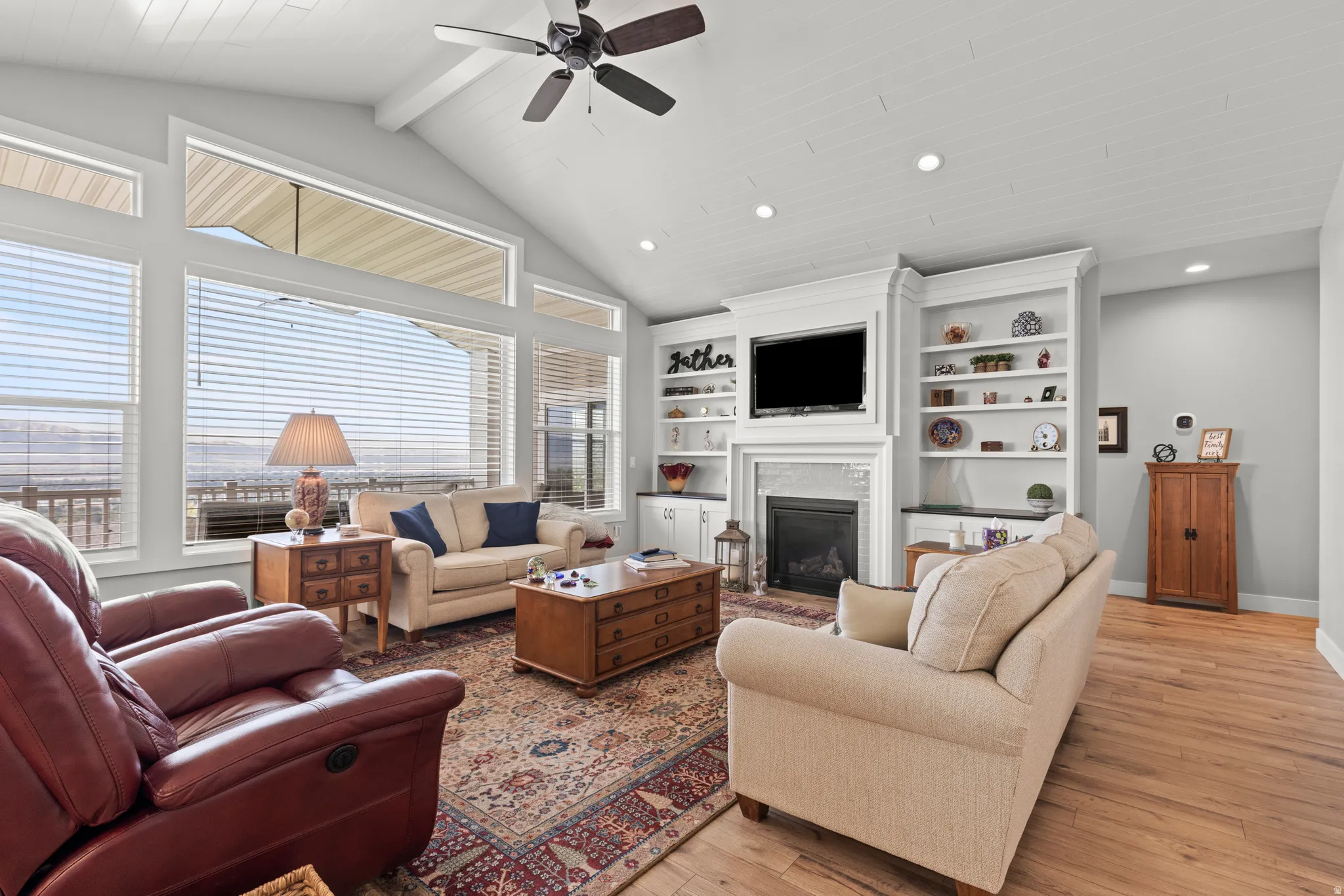 Vaulted ceiling living area featuring a fireplace with stone surround, custom built-in shelving, wood-finish flooring, and expansive windows