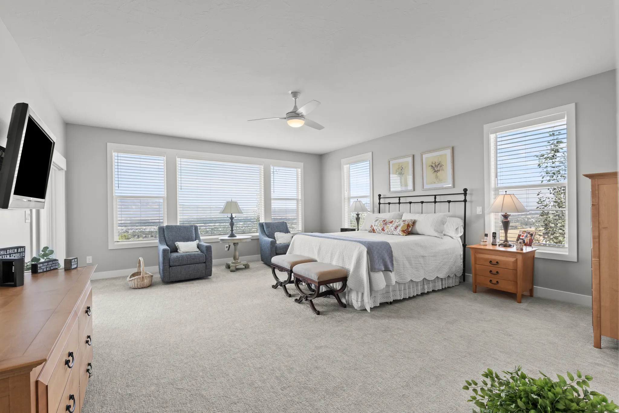 Spacious sleeping area featuring light gray wall paint, light-colored carpeting, multiple windows with blinds, and a ceiling fan with integrated lighting