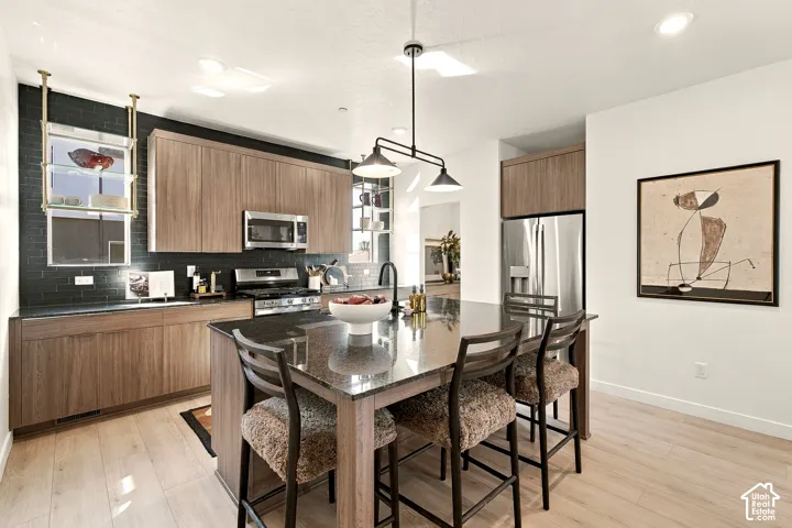 Kitchen featuring flat-panel wood-finish cabinetry, black subway tile backsplash, dark stone-surface island with integrated seating, stainless steel appliances, and matte black faucet fixtures