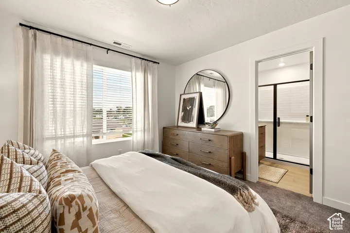 Bedroom featuring neutral wall tones, plush carpeting, a wide window with black hardware, and an entryway to a bathroom with a glass-enclosed shower