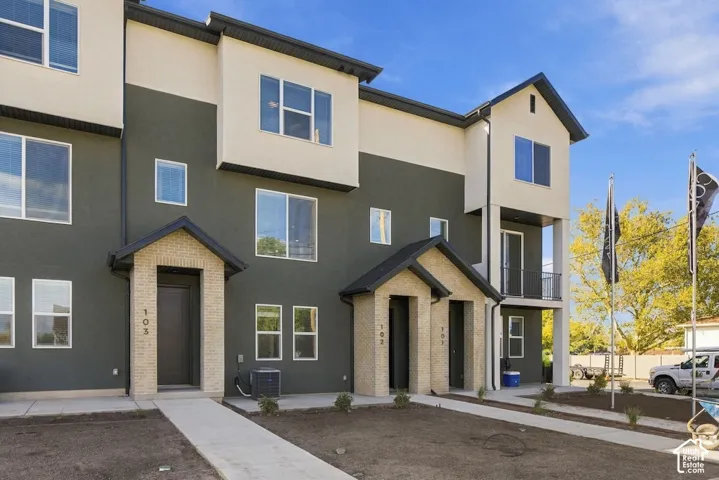 Three-story multi-unit exterior featuring dual-tone stucco siding, light brick entrance detailing, covered porches, and a second-level balcony with metal railing