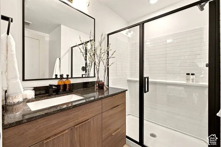 Bathroom featuring a wood-finish vanity with a dark stone countertop, undermount sink, black-framed mirror, and a walk-in shower with white subway tile and a black metal frame