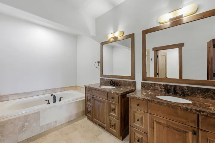 Bathroom featuring a built-in soaking tub with stone tile surround, dual wood vanities with integrated sinks, dark stone countertops, and framed mirrors with overhead lighting