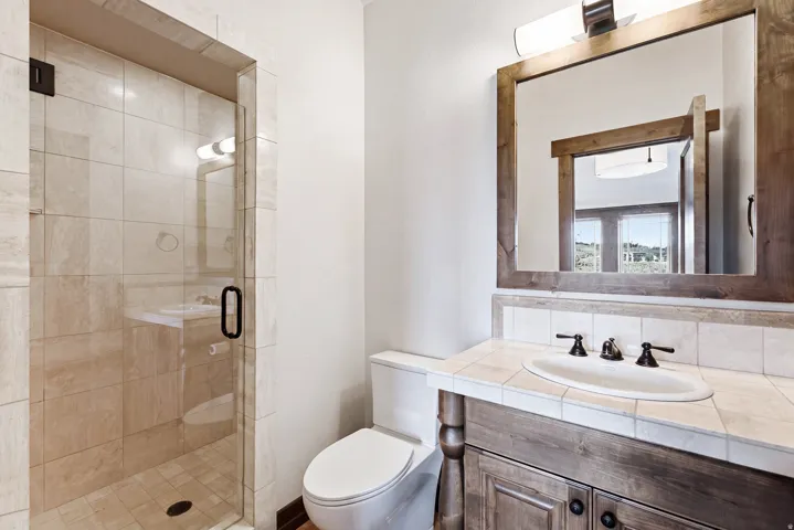 Bathroom featuring a walk-in shower with clear glass enclosure and tile surround, a built-in wood-finish vanity with an undermount sink, tile countertop, and a wood-framed mirror