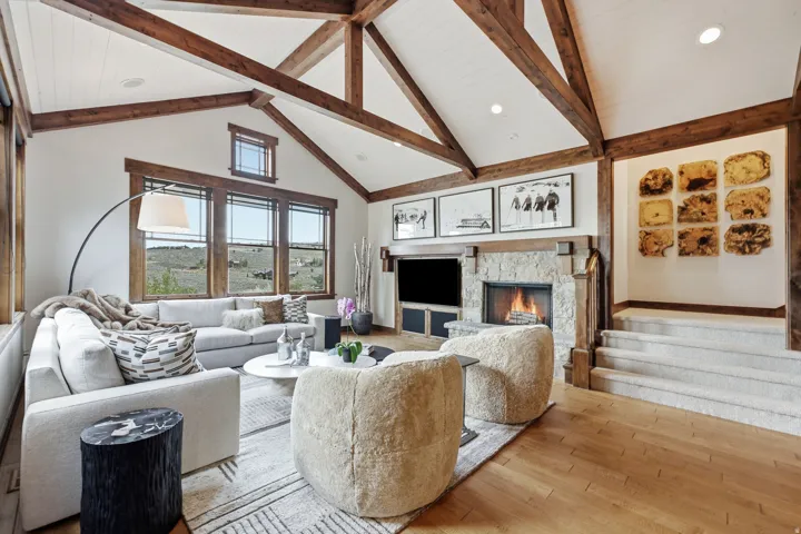 Vaulted ceiling with exposed wood beams, stone fireplace, wood-finish flooring, and multiple windows with wood trim