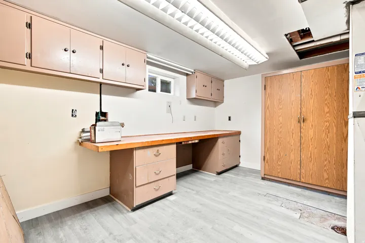 Utility room featuring extensive built-in cabinetry, a dedicated workbench with integrated drawers, wood-finish flooring, a fluorescent ceiling light fixture, and a single window