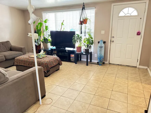 Spacious room featuring neutral tile flooring, a white entry door with an arched window insert, and a large window with horizontal blinds