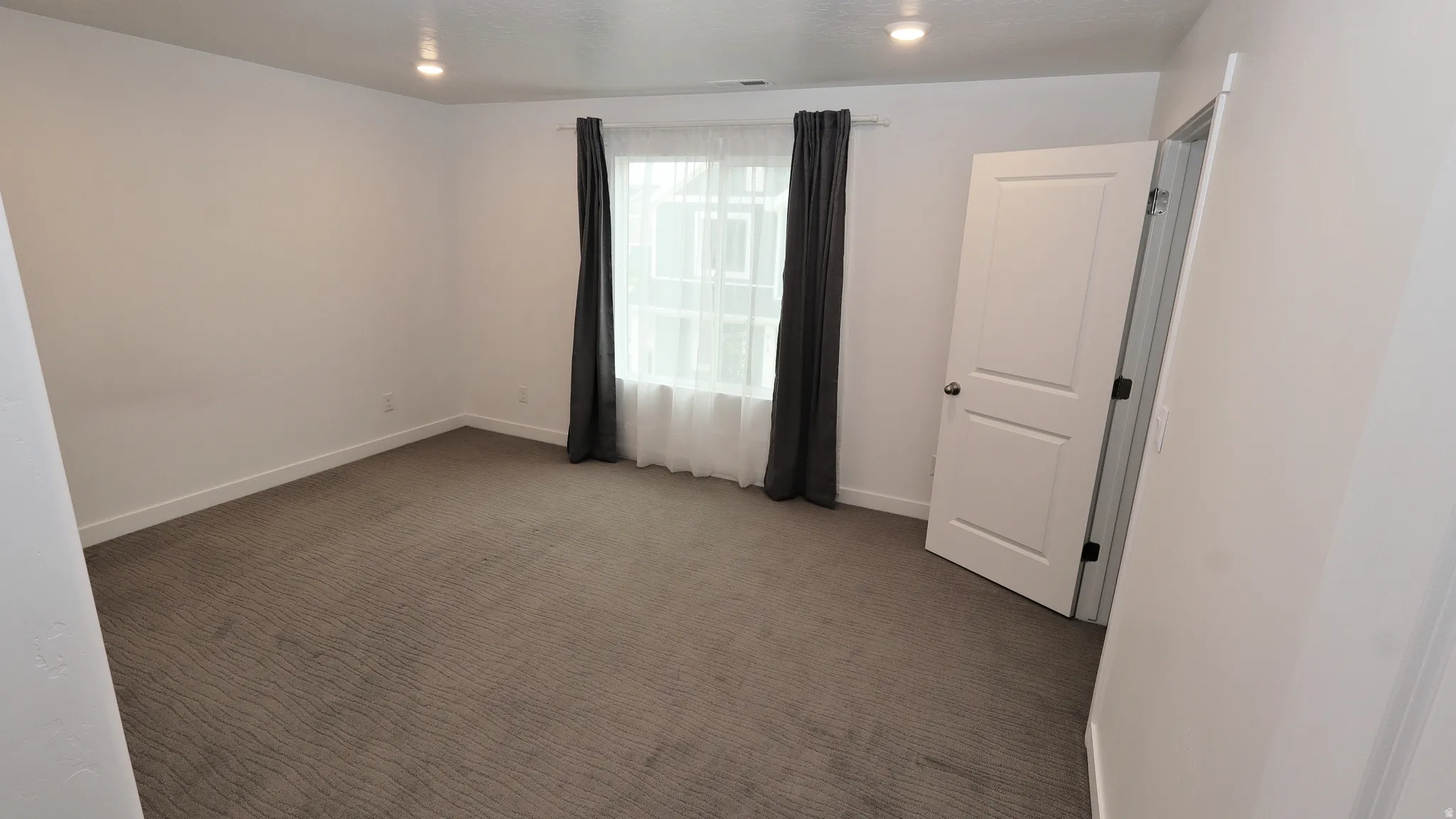Spacious room featuring neutral wall tones, patterned carpet flooring, a window with sheer and blackout curtains, recessed ceiling lighting, and a paneled interior door with dark hardware