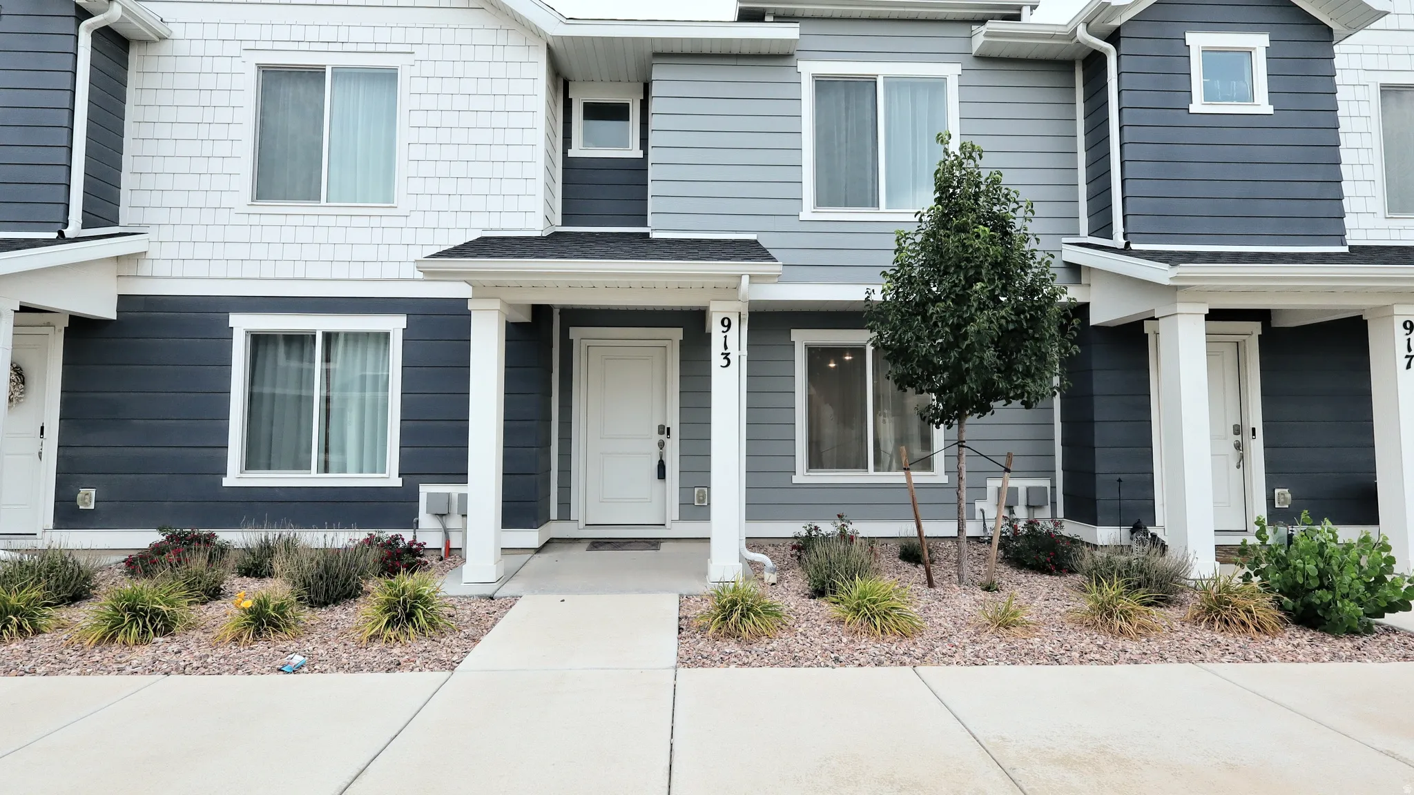Townhome exterior featuring varied siding in shades of blue and white, a private entrance, and a well-maintained landscaping bed with a young tree