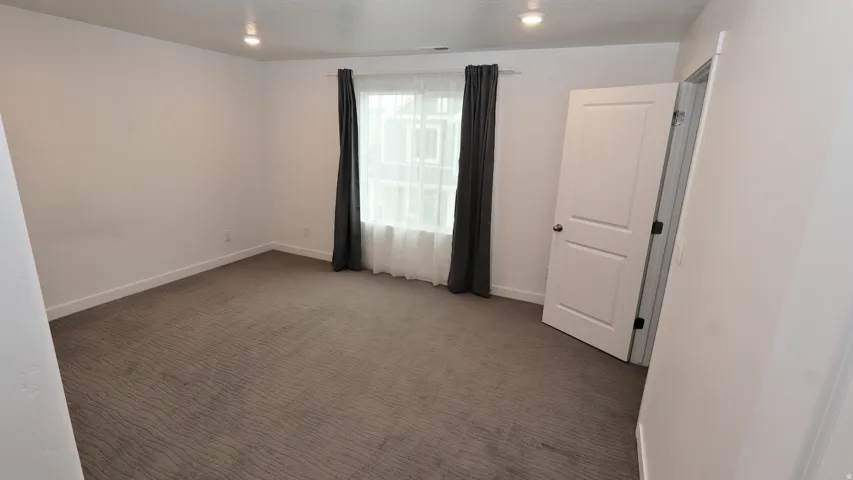 Spacious room featuring neutral wall tones, patterned carpet flooring, a window with sheer and blackout curtains, recessed ceiling lighting, and a paneled interior door with dark hardware