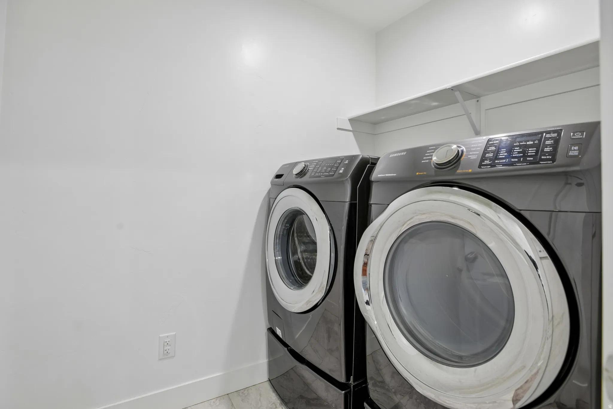 Dedicated laundry area featuring a front-loading washer and dryer, an overhead storage shelf, and light-toned flooring