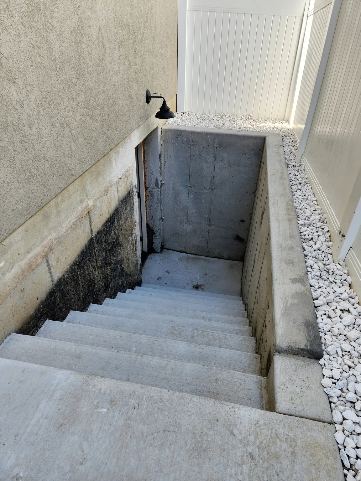 Exterior concrete staircase leading to a lower-level entrance, featuring a stucco-finish wall, a black wall-mounted light fixture, and white gravel landscaping bordering a white fence