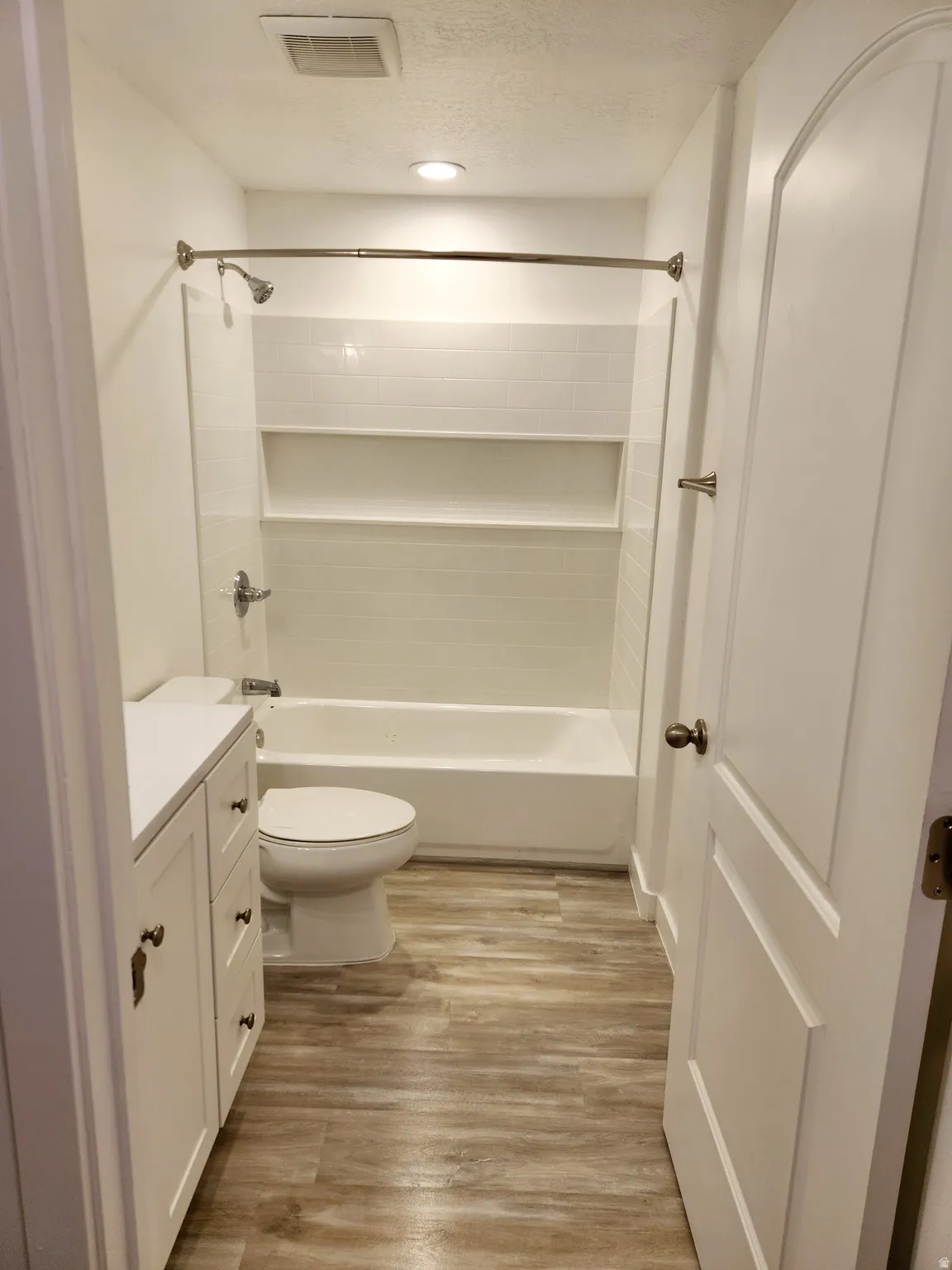 Bathroom featuring a full-size bathtub with subway tile surround and integrated shelving, wood-finish flooring, a white vanity with cabinet storage, and a paneled door