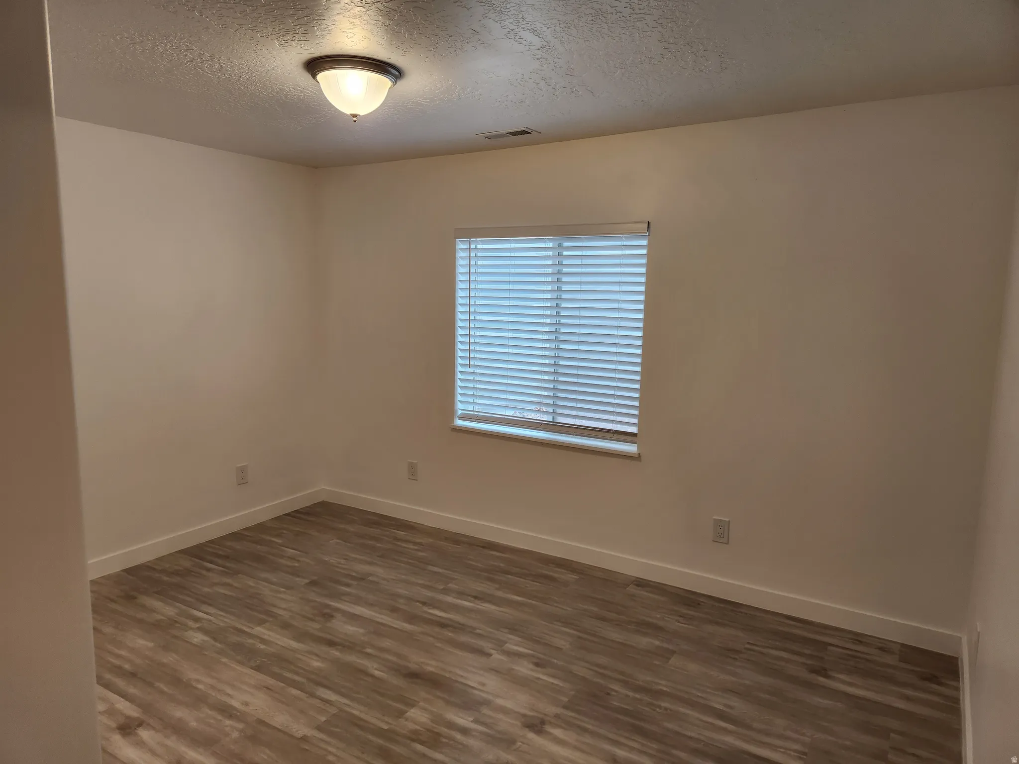 Room featuring wood-finish flooring, a window with blinds, a flush-mount ceiling light fixture, and neutral wall tones