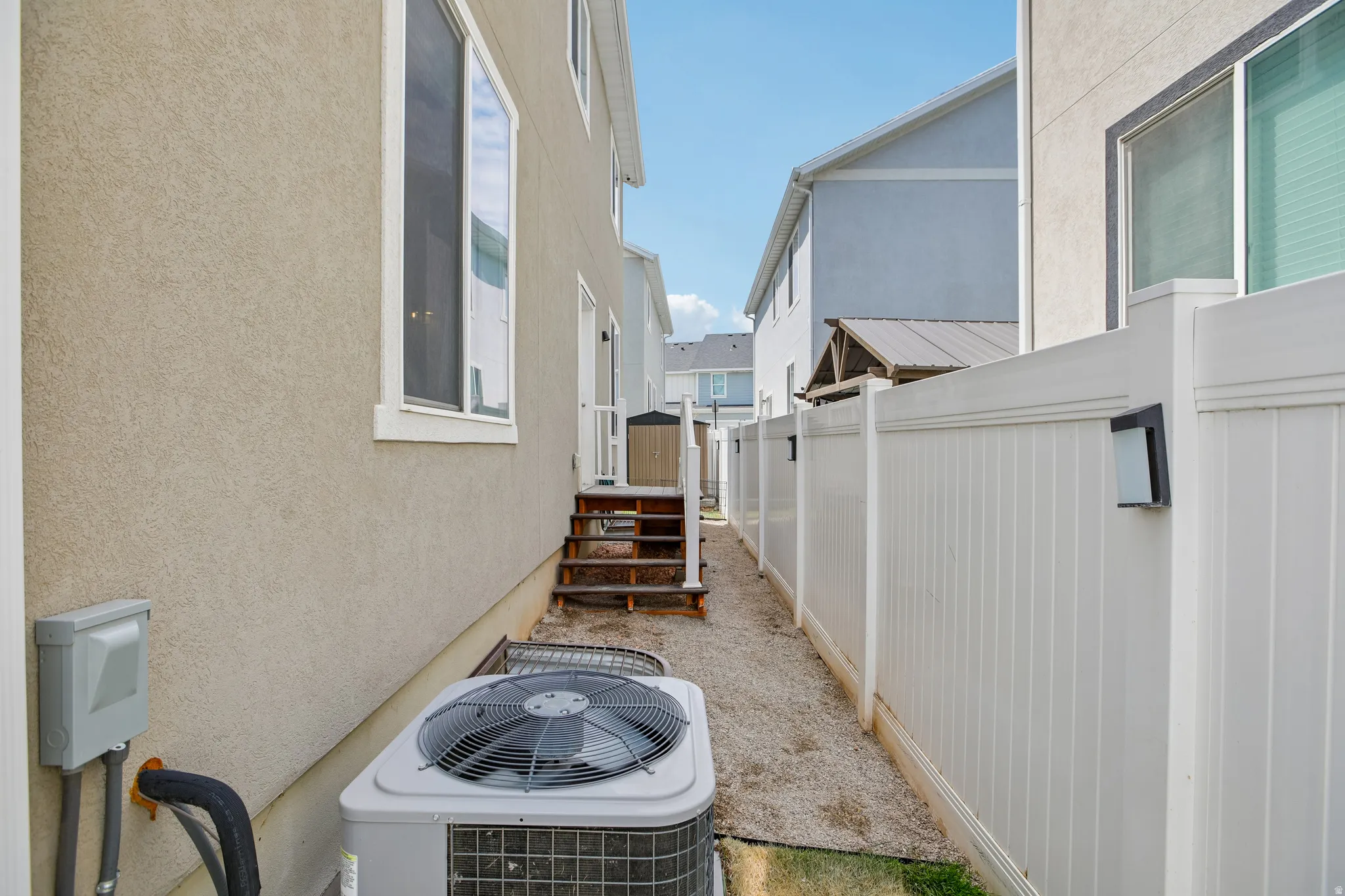 Side yard with an air conditioning unit, stucco exterior, white vinyl fencing, and wooden stairs leading to an upper-level entry