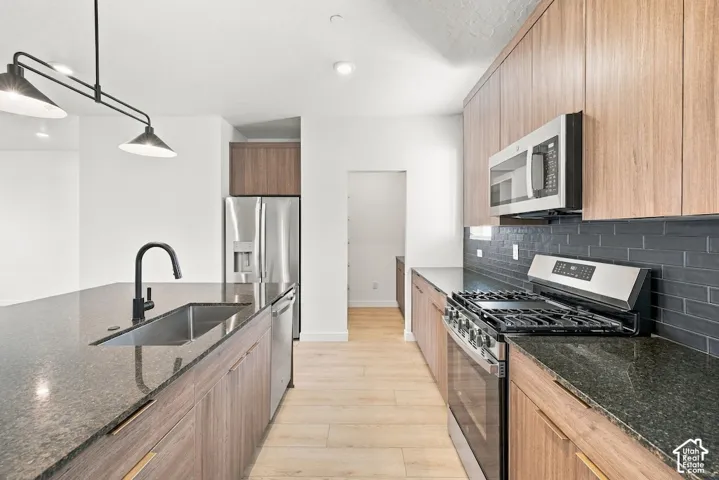 Kitchen featuring wood-finish cabinetry, dark stone countertops, stainless steel appliances, a dark tile backsplash, and a matte black gooseneck faucet