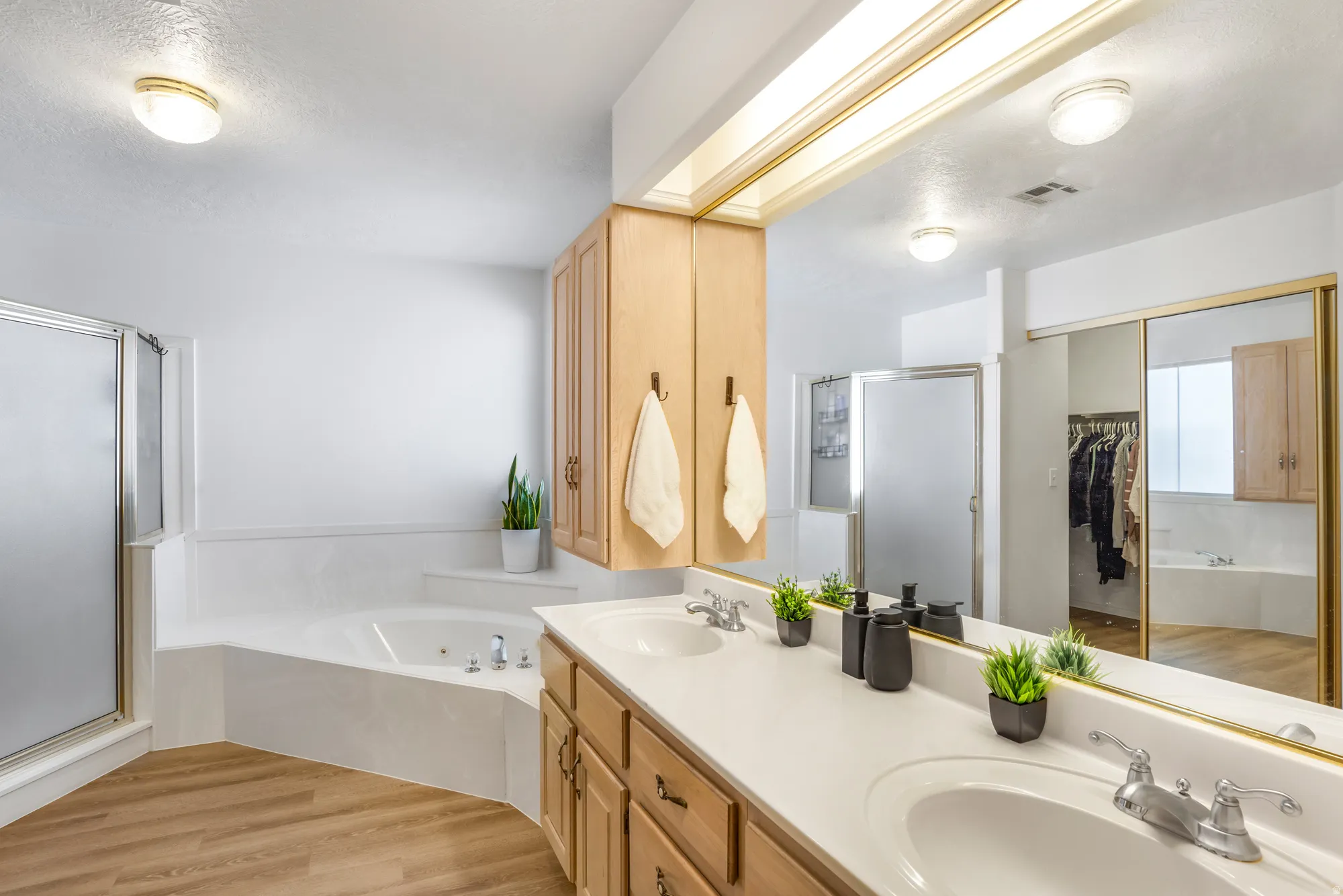 Bathroom featuring a dual vanity with light-colored countertops, a large framed mirror, built-in wooden cabinetry, a corner jetted tub, and a separate shower enclosure with a frosted glass door