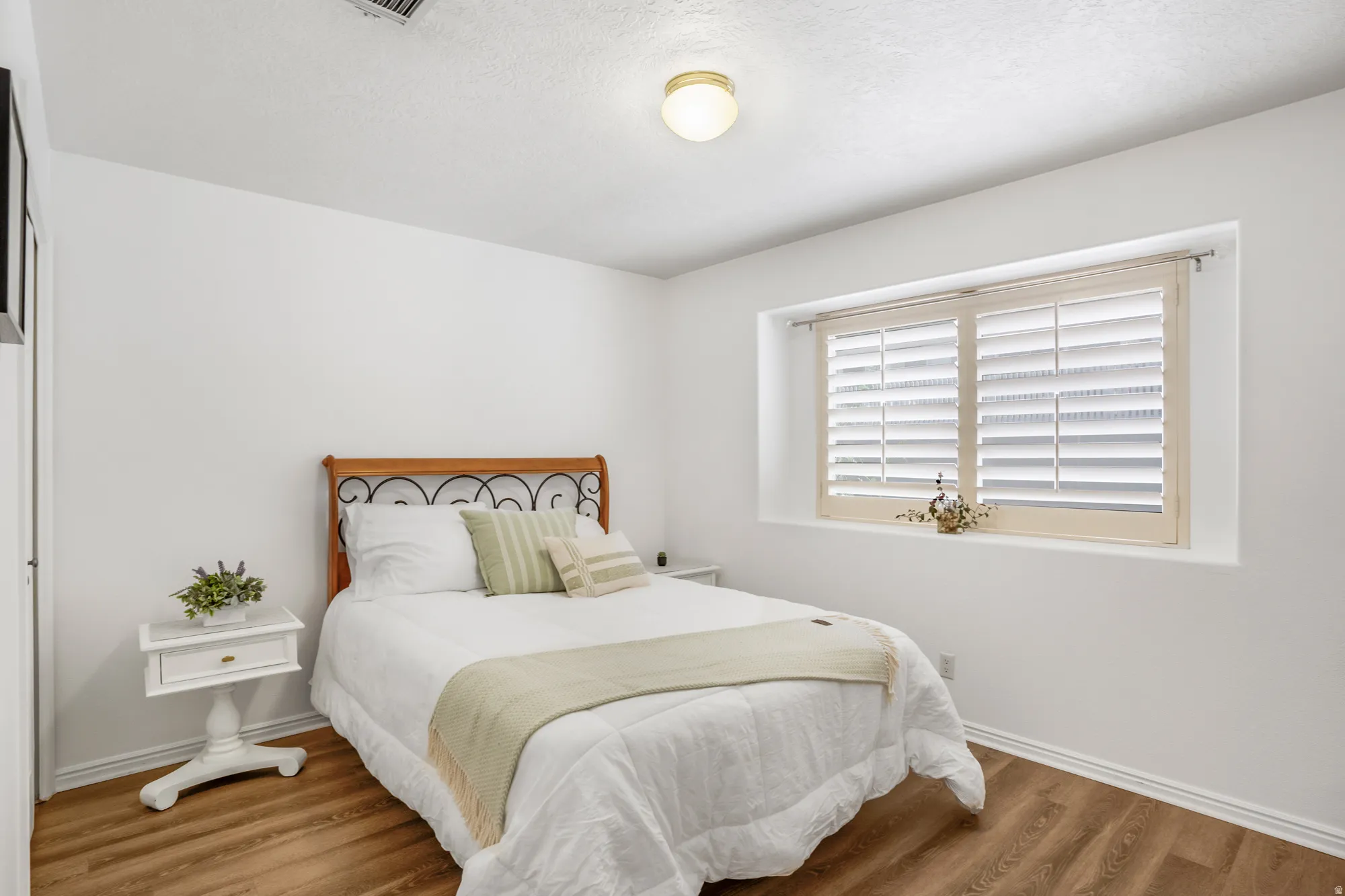 Bedroom featuring wood-finish flooring, a window with plantation shutters, white walls, and a ceiling-mounted light fixture