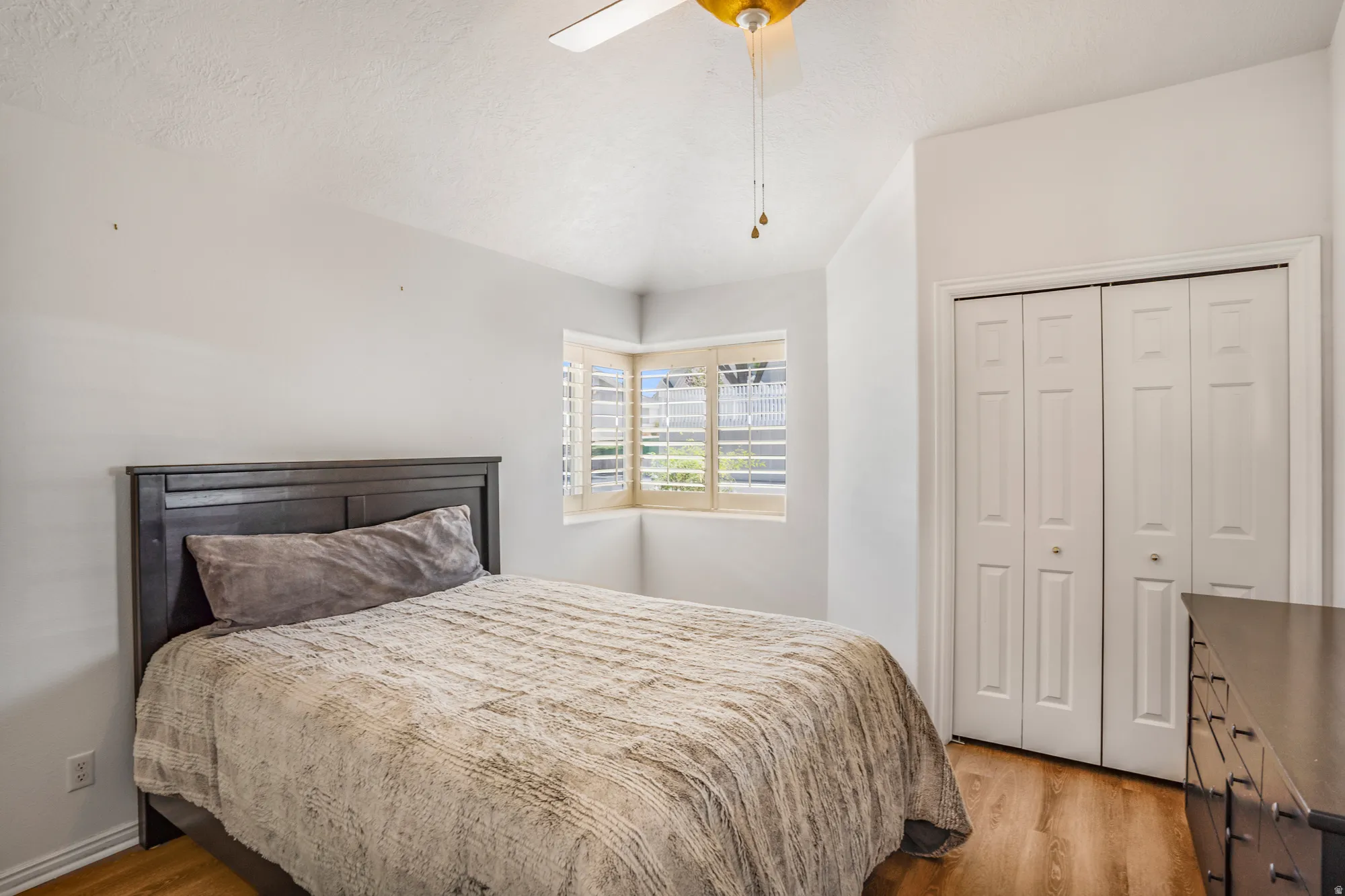 Bedroom with wood-finish flooring, corner windows with plantation shutters, bifold closet doors, and a ceiling fan with light fixture