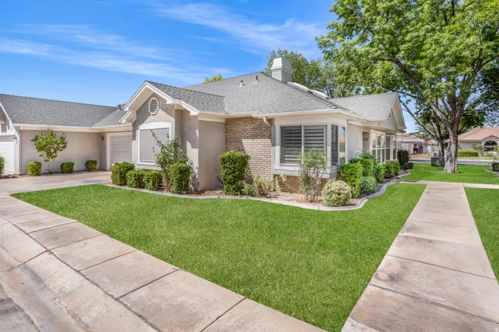 Single-story residence featuring a stucco and brick exterior, a grey shingle roof, bay window with plantation shutters, attached two-car garage, and a manicured front lawn with mature landscaping