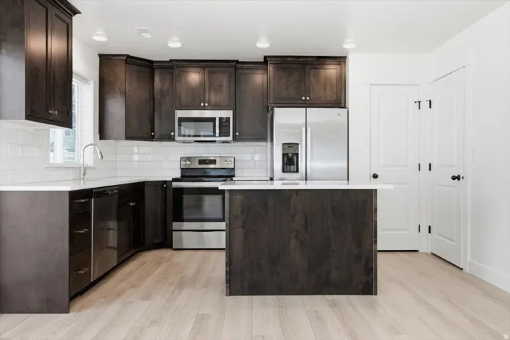 Kitchen featuring dark wood-finish cabinetry, stainless steel appliances, white subway tile backsplash, light wood-finish flooring, and a central island with white countertop
