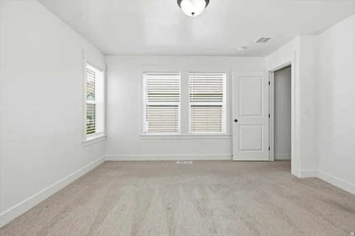 Spacious room featuring light gray carpeting, crisp white walls, two large windows with blinds, a white paneled door with black hardware, and a semi-flush mount ceiling light fixture