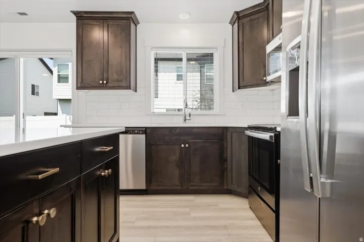 Contemporary kitchen featuring dark wood cabinetry, light-toned wood-finish flooring, white subway tile backsplash, and stainless steel appliances including a refrigerator, dishwasher, and range