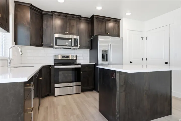 Modern kitchen featuring dark wood-finish cabinetry, stainless steel appliances, white subway tile backsplash, light wood-finish flooring, and a central island with a white countertop