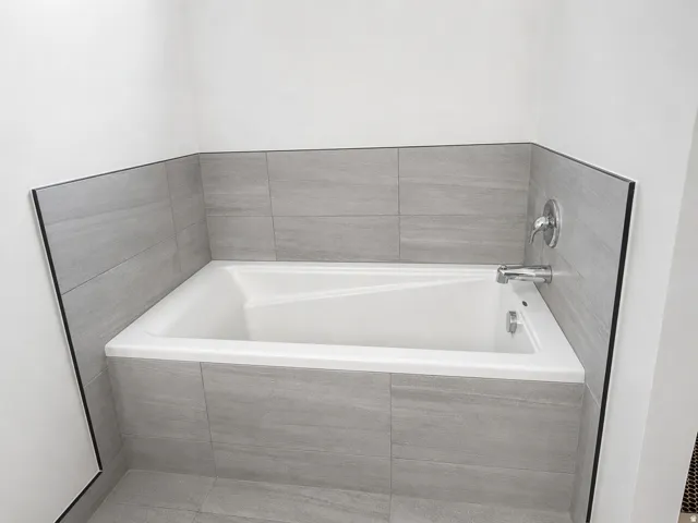 Built-in bathtub with a white finish, surrounded by light gray tiled walls and a coordinating tiled tub surround