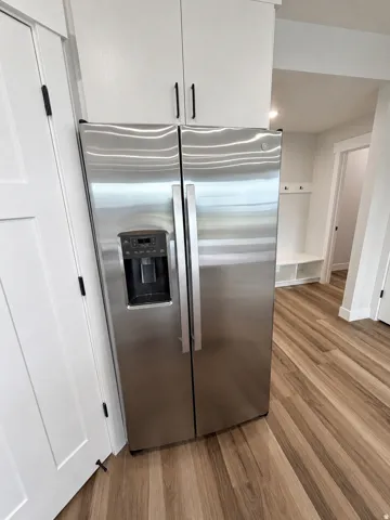 Stainless steel side-by-side refrigerator, white upper cabinetry with matte black hardware, and wood-finish flooring