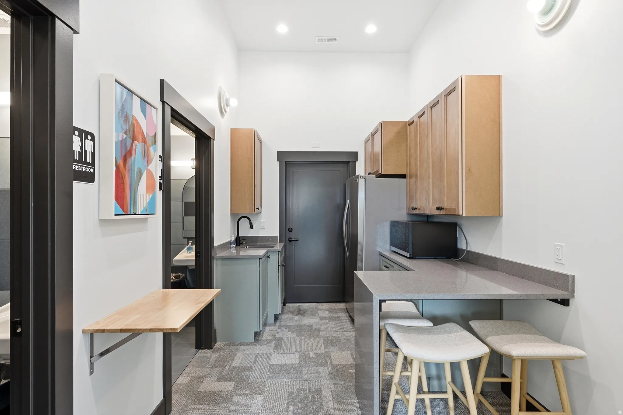 Compact kitchen with light wood-finish cabinetry, a black faucet, and a grey countertop extending to a breakfast bar