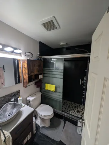 Bathroom featuring a stone vessel sink, dark wood vanity, stone countertop, toilet, and a shower with a sliding glass door and pebble tile floor