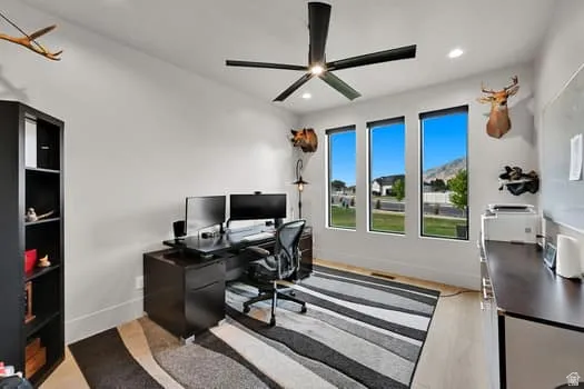 Bright room featuring three vertical windows, a modern black ceiling fan, recessed lighting, light wood-finish flooring, and white walls