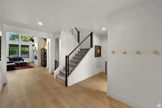 Open-concept living space featuring light wood-finish flooring, a modern staircase with black railings and cable infill, a stone fireplace, and large windows with transom details