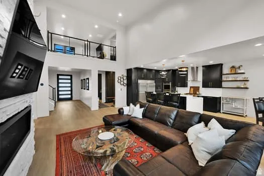 Open-concept main living area featuring wood-finish flooring, a contemporary fireplace with a stacked stone facade, a kitchen with dark cabinetry and stainless steel appliances, and a second-story interior balcony with black railings
