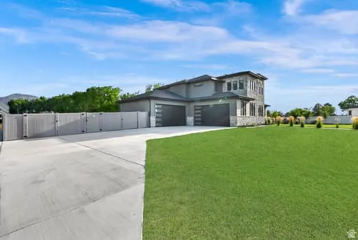 Modern residence featuring a stone and stucco exterior, a manicured lawn, an expansive concrete driveway, a multi-panel perimeter fence, and two contemporary garage doors with frosted glass accents