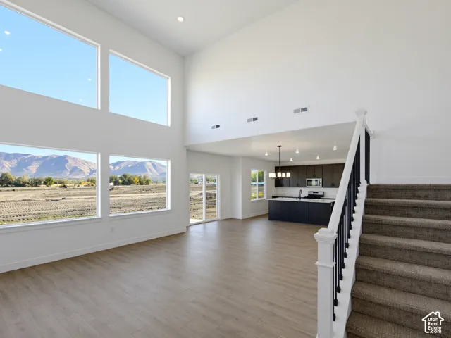 Expansive double-height ceilings, floor-to-ceiling windows featuring mountain scenery, wood-finish flooring, kitchen island with built-in cabinetry, and staircase with white railings and dark iron balusters