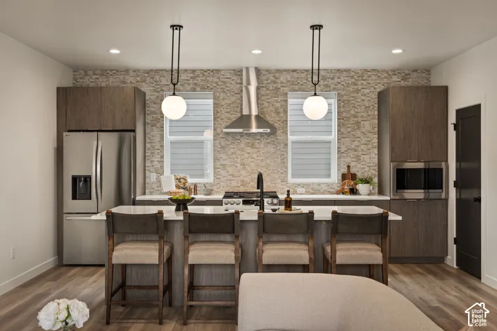 Kitchen featuring wood-finish cabinetry, a textured stone backsplash, quartz-topped island with seating, stainless steel appliances, and dual pendant light fixtures