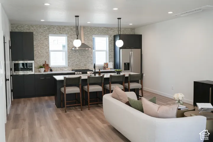 Kitchen featuring dark cabinetry, quartz-topped center island, stacked stone backsplash, stainless steel range hood, and wood-finish flooring