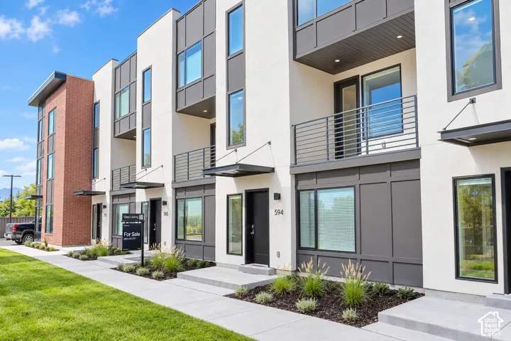 Modern three-story exterior featuring a mix of stucco and brick siding, expansive vertical windows, private metal-railed balconies, and contemporary cantilevered entry awnings