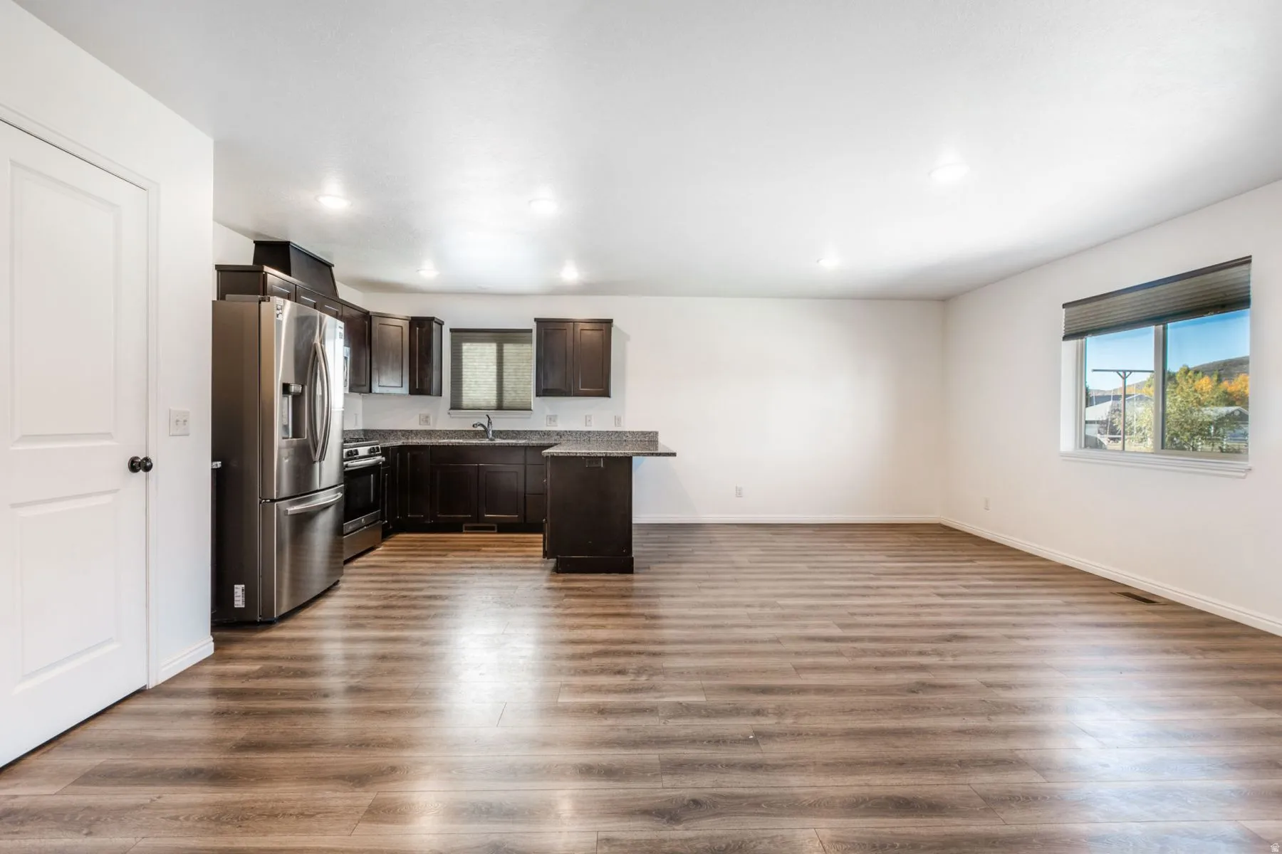 Open-concept living area with wood-finish flooring, recessed lighting, and a kitchen featuring dark cabinetry, stainless steel appliances, and a breakfast bar