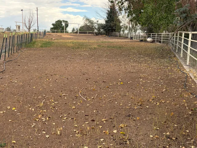 Expansive open lot featuring a white pipe fence, a section of black T-post fencing, mature trees, and a visible propane tank