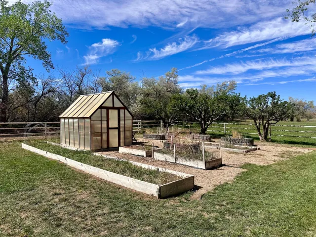 Greenhouse structure, multiple raised garden beds, established trees, and a wooden perimeter fence